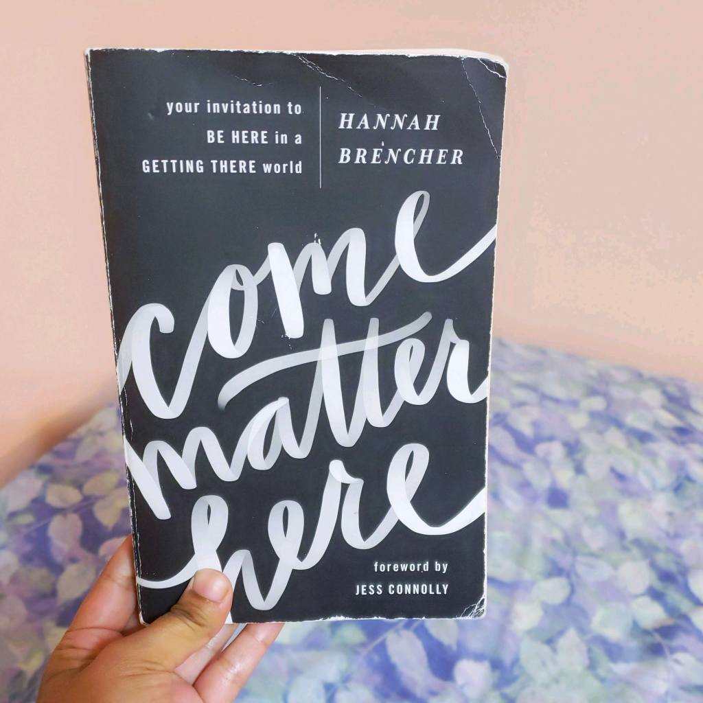 A brown hand holds up a worn copy of Come Matter Here by Hannah Brencher up against a background of a pink wall and a bed with a light blue and purple sheet over it.