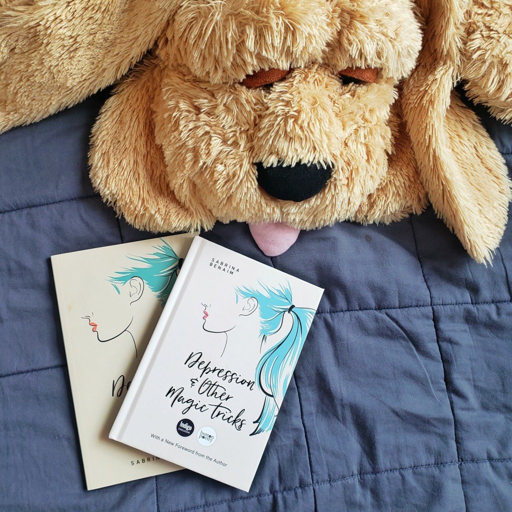 Two copies of depression and other magic tricks by Sabrina Benaim are laid out on top of a gray blanket. One is a paperback and the other is the hardcover edition. There is a yellow stuffed dog lying near the two copies.