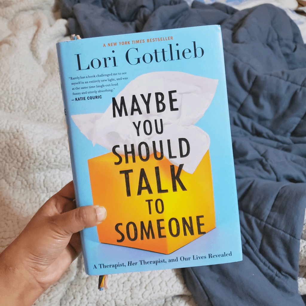 A brown hand holds up a copy of maybe should talk to someone over a bed with the couple blankets laid out 