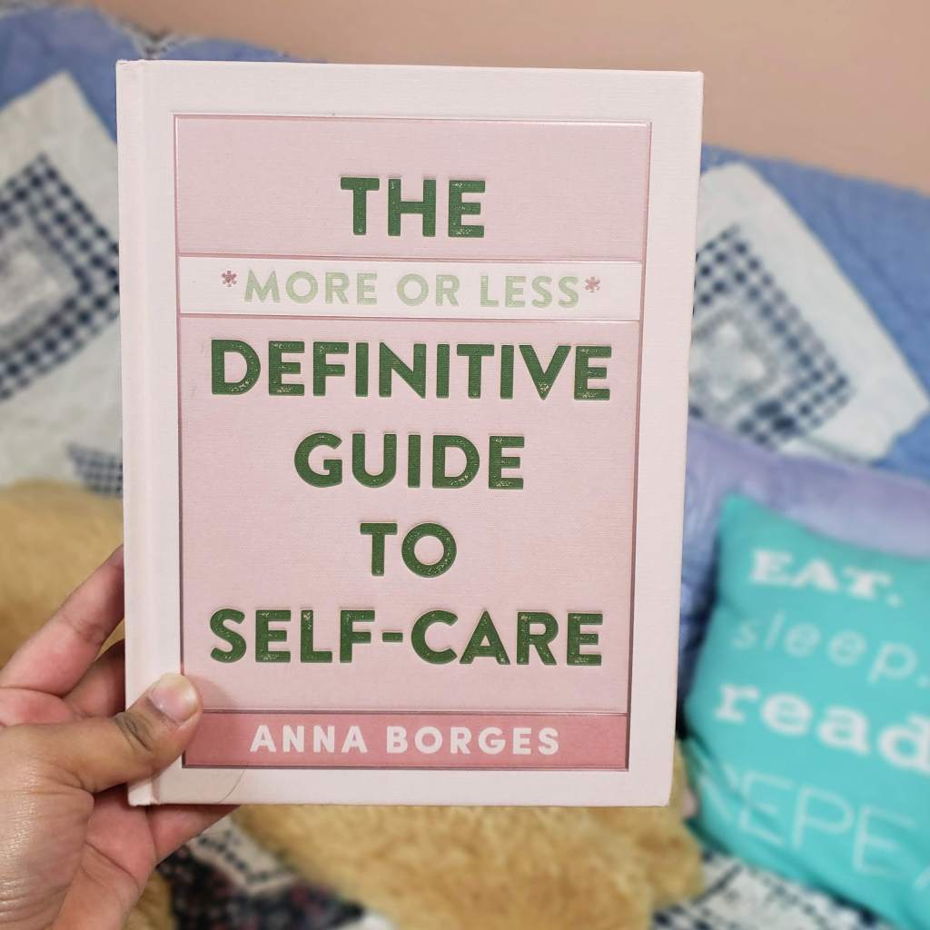 A brown hands holds up a copy of the more or less definitive guide to self care by anna borges 